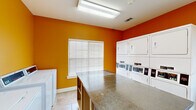 This self-serve laundry facility at 3300 Stadium Dr in Phenix City, Alabama, is a compact yet efficiently organized space designed to meet residents' laundry needs around the clock. The room is immediately recognizable by its bold, warm orange walls, which lend an energetic and welcoming ambiance to an otherwise utilitarian space. Bright fluorescent overhead lighting ensures excellent visibility throughout the room at any hour. The facility is equipped with multiple stacked commercial dryers and top-load washing machines with coin-operated controls, arranged along the walls to maximize usable floor space. A large granite-topped folding island at the center of the room provides generous counter space for sorting and folding laundry. Light-toned tile flooring runs throughout, offering a clean, easy-to-maintain surface. A window fitted with white horizontal blinds allows natural light to filter in, brightening the interior during daytime hours. An exterior-facing glass door provides direct outdoor access, further enhancing ventilation and convenience. Additional open shelving units along one wall offer supplemental storage space. A glass interior door connects the laundry room to an adjacent fitness center, indicating this facility is part of a broader community amenity complex. Safety features include a clearly marked exit sign and fire extinguisher.