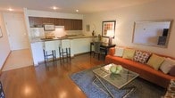This single-floor space is designed around an open-concept layout that connects the kitchen, dining, and living areas into one cohesive and functional environment. Warm hardwood flooring flows throughout the entire space, lending a sense of continuity and warmth to the interior.

The kitchen is positioned along one wall and features rich dark wood cabinetry, a built-in microwave, a stainless steel sink, and stone-look countertops. A peninsula counter with bar seating provides a convenient space for casual dining or meal prep, maximizing the utility of the layout.

Adjacent to the kitchen, a compact dining area offers an intimate setting for meals, while the living area anchors the space with a bold orange sofa, a glass-top coffee table, and a plush area rug. Recessed lighting in the ceiling provides clean, even illumination throughout the space.

The overall ambiance is modern yet inviting, with a carefully curated mix of warm tones, natural materials, and contemporary furnishings. The open floor plan enhances the sense of space, making the interior feel airy and well-organized. Tasteful décor accents, including framed artwork and decorative lighting, add personality and a welcoming character to the space.