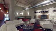The Phase I Resident Lounge is a dynamic, industrial-style communal space designed to offer residents a well-appointed retreat for socializing and relaxation. The lounge's open-concept layout is anchored by soaring ceilings with exposed ductwork, polished concrete floors, and a striking mezzanine level accessible via a sleek metal staircase with railing, adding a dramatic vertical dimension to the space.

The kitchenette area features rich wood cabinetry, a green-accented bar counter with modern bar stools, and a stainless steel refrigerator — ideal for casual gatherings. Bold, swirling area rugs define each zone and inject vibrant color throughout.

The lounge area is furnished with plush white sofas and armchairs arranged around a glass-top coffee table, creating an inviting seating arrangement. A full-height stacked stone fireplace with built-in shelving serves as a commanding focal point, complemented by a wall-mounted flat-screen television nearby. A billiards table set beneath pendant lighting adds an entertainment-focused element to the space.

Exposed pipes, structural columns, and industrial-style pendant fixtures reinforce the loft-inspired aesthetic, while wall sconces and warm ambient lighting throughout contribute to an inviting, social atmosphere. Framed artwork on the walls adds a curated finishing touch to this well-designed resident amenity.