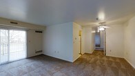 This single-floor residence offers a straightforward and functional layout designed for comfortable everyday living. The main living area features wall-to-wall beige carpeting that extends throughout, providing a warm and cohesive feel underfoot. Clean white walls and smooth ceilings create a bright, neutral backdrop throughout the space.

A partial dividing wall separates the living area from the adjacent kitchen zone, which is visible in the background and includes a refrigerator and what appears to be additional cabinetry. This separation provides a sense of definition between living and cooking areas while maintaining an open sightline.

A hallway extends from the main living space toward the back of the residence, illuminated by a ceiling fan with an integrated light fixture — a feature that also appears in at least one additional room visible along the corridor. The hallway leads to what appear to be one or more private rooms, suggesting a multi-room configuration.

Ventilation is handled through floor-level baseboard registers and ceiling vents, indicating a central HVAC system. The overall design is clean and utilitarian, with practical finishes throughout. The residence is presented in a vacant state, offering a clear view of its layout, proportions, and structural features without furnishings as a distraction.