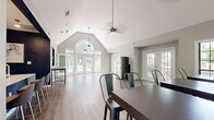 This single-floor community amenity facility offers a thoughtfully designed layout encompassing a clubroom, a kitchenette area, and a dedicated fitness center. The clubroom is an architectural standout, featuring dramatic vaulted ceilings with an arched transom window that floods the space with natural light. Large multi-pane windows line the walls, framing views of surrounding mature trees. The room is furnished with dark wood communal tables and industrial-style metal chairs, complemented by light hardwood-style flooring that runs throughout. A ceiling fan with pendant lighting adds to the airy, open ambiance.

The kitchenette area is sleekly appointed with a bold navy blue island trimmed in white, metallic tile backsplash, and bar seating for casual gatherings. A full-size refrigerator and ample counter space make it well-suited for events. A herringbone-patterned accent wall adds a refined decorative touch.

The fitness center is energetically styled with vivid blue and green accent walls, vaulted ceilings, and ceiling fans. It is comprehensively equipped with treadmills, a rowing machine, cable machines, a Smith machine, free weights, benches, stability balls, and wall-mounted televisions. Multiple French doors and large windows ensure the gym remains bright and connected to the outdoors throughout the day.