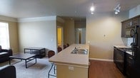 This single-floor space is designed around an open-concept layout that seamlessly connects the kitchen and living areas. The kitchen features a peninsula with a double-basin sink and laminate countertop, providing both a functional workspace and a casual dining area with seating for several bar-style chairs. The flooring transitions from hardwood-style material in the kitchen zone to plush carpet in the living area, clearly defining each functional space.

The living area is modestly sized, accommodating a small desk and shelving unit, and benefits from the open sightlines created by the connected layout. Track lighting is installed in the kitchen area, providing focused illumination. The walls are finished in a warm neutral tone throughout, contributing to a cohesive and understated aesthetic.

Doorways visible in the background lead to additional rooms, including what appears to be a bathroom with warm lighting and a secondary space, suggesting a functional multi-room floor plan. Crown molding details near the ceiling add a subtle architectural accent. Overall, the space presents a practical, straightforward layout suited for everyday living.