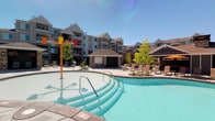 This outdoor amenity space is a well-appointed resort-style recreation area designed for relaxation and entertainment. At the heart of the space is a spacious swimming pool with clear turquoise water, featuring a gradual entry and a colorful splash pad area, making it ideal for residents of all ages. The pool deck is generously lined with lounge chairs, offering plenty of room to unwind under the open sky.

A charming covered pavilion anchored in stone serves as a central gathering point, providing shade and a focal area for outdoor socializing. The pavilion is complemented by additional seating arrangements nearby, creating a comfortable and inviting atmosphere throughout the space.

Mature trees and thoughtfully placed landscaping add a natural element that softens the hardscape and enhances the overall aesthetic. The surrounding multi-story residential buildings feature balconies and neutral-toned exteriors with pitched rooflines, framing the amenity area with an architectural backdrop that blends contemporary and traditional design elements.

The space benefits from abundant natural light and open sky views, creating a bright and energetic environment. Whether lounging poolside, enjoying the splash features, or gathering under the pavilion, this outdoor amenity area offers a comprehensive and appealing recreational experience.