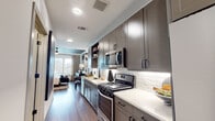 This well-appointed single-floor residence combines modern design with thoughtful functionality across its open layout. The galley-style kitchen is a standout feature, outfitted with grey shaker-style cabinetry, crisp white quartz countertops, a white subway tile backsplash, and a full suite of stainless steel appliances including a gas range, over-the-range microwave, and refrigerator. A matte black pull-down faucet over a deep single-basin sink adds a sleek, contemporary touch, while under-cabinet lighting enhances both form and function. Open shelving provides additional display and storage space. The kitchen flows naturally toward the living area, which benefits from natural light filtering in through large windows. The bedroom is a serene retreat defined by a rich teal accent ceiling, wide windows with outdoor views, warm wood-tone flooring, and a ceiling fan. A double-door closet and built-in shelving provide ample storage, while a cozy reading nook with decorative gold-framed mirrors adds character. The bathroom continues the modern aesthetic with large-format grey tile flooring, a glass-enclosed walk-in shower, hexagonal tile wall art, and matte black hardware accents. Wood-look flooring runs consistently throughout the living spaces, unifying the home's cohesive, contemporary design.