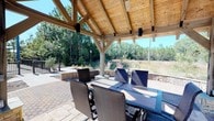 This single-level outdoor pavilion is a masterfully designed al fresco entertaining space, showcasing a striking vaulted ceiling constructed from warm, natural cedar wood with exposed timber framing. The open-air structure allows abundant natural light to filter through while providing ample shade and shelter for outdoor gatherings. A decorative ceiling fan is centrally mounted to ensure comfortable airflow, and built-in wall speakers are positioned throughout the pavilion, making it ideal for entertaining.

The space is furnished with a large rectangular dining table paired with wicker-woven chairs, comfortably seating multiple guests for outdoor dining. The flooring consists of beautifully laid paver stones, adding a refined, durable surface underfoot. Adjacent to the pavilion, a raised stone-accented planter or water feature adds architectural interest and visual depth to the patio area.

The pavilion opens fully to panoramic views of lush, natural greenery, including tall pines and native grasses that create a serene, private backdrop. A paved walkway and landscaped garden beds extend beyond the pavilion, enhancing the overall outdoor living experience. The combination of natural wood, stone accents, and surrounding woodland creates a harmonious and inviting atmosphere perfectly suited for relaxation and entertaining.