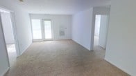 This single-floor space presents a straightforward and functional layout centered around an open living area. The room is finished with wall-to-wall neutral beige carpeting that extends throughout, creating a cohesive and understated foundation. White walls keep the interior bright and versatile, providing a blank canvas for a variety of interior design approaches.

A pair of white-framed glass doors along one wall allows natural light to filter into the space, contributing to an airy atmosphere. A wall-mounted HVAC vent is positioned nearby, indicating a built-in climate control system. The open layout of the main living area offers flexibility in furniture arrangement and use of the space.

A doorway visible along one wall leads to an adjacent room, suggesting additional living or functional areas beyond the main space. The transition between rooms is defined by clean white door framing that maintains the consistent, neutral aesthetic found throughout. The overall ambiance is clean, practical, and move-in ready, suited for a range of occupants seeking a simple, unadorned living environment.