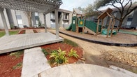 This outdoor community space is thoughtfully designed to offer a variety of recreational amenities within a well-maintained courtyard setting. At its center stands a wooden pergola deck with string lighting, accessed via a concrete pathway flanked by mulched planting beds with ornamental foliage. The pergola provides a shaded gathering area ideal for relaxation or socializing.

Adjacent to the pergola, a children's play structure features green and wood-toned components including slides, climbing panels, and interactive elements, all set on a gravel safety surface bordered by rubber edging. A separate open lawn area hosts a stone fire pit with surrounding bench seating, providing an inviting space for evening gatherings beneath mature shade trees.

Two substantial outdoor grilling stations, finished in stacked stone tile with granite-style countertops, anchor the grilling area and offer ample prep space for outdoor cooking. Concrete pathways connect the various activity zones throughout the courtyard, ensuring easy navigation. Red mulch landscaping, mature trees, and greenery contribute to the overall ambiance, creating a functional and welcoming outdoor environment.