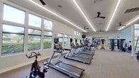 This modern fitness center is thoughtfully designed to support a comprehensive workout experience across its single-floor layout. The space is bathed in natural light courtesy of expansive floor-to-ceiling windows that line multiple walls, offering pleasant outdoor views of surrounding greenery. Linear LED lighting runs along the ceiling, complemented by ceiling fans that maintain airflow and comfort throughout the facility.

The cardio zone features a generous row of Matrix treadmills, elliptical trainers, and stationary cycling bikes, all positioned to face the windows — making workouts feel open and energizing. A separate strength training area houses a multi-functional cable machine, free weights, benches, medicine balls, resistance equipment, and various accessories for functional fitness training. Large wall-to-wall mirrors in the strength zone enhance the sense of space while allowing for proper form checks during exercise.

The flooring throughout is a stylish multi-toned carpet tile with distinctive orange accent stripes, adding a contemporary visual element to the space. Wall-mounted flat-screen TVs are positioned throughout the facility for entertainment during workouts. A glass-paneled entry door provides easy access to an adjacent outdoor area. The overall ambiance is bright, clean, and professional, making this fitness center an inviting and functional destination for a wide range of exercise routines.