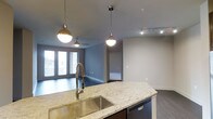 This single-floor Dallas residence showcases a thoughtfully designed open-concept layout that seamlessly connects the kitchen and living areas. The kitchen is a standout feature, centered around a curved granite island countertop with warm tonal variations, paired with a professional-style stainless steel pull-down faucet and an undermount sink. A dishwasher is integrated into the island cabinetry, adding practicality to the sleek design. Overhead, globe-style pendant lights hang from polished chrome chain fixtures, complementing the track lighting that runs along the ceiling of the adjoining living space.

The living area benefits from natural light streaming through a set of large windows fitted with blinds, offering both brightness and privacy. A ceiling fan is positioned centrally in this zone, contributing to comfort year-round. Dark hardwood-style flooring flows throughout, providing a rich contrast to the light gray walls. The neutral color palette and clean architectural lines create a modern, cohesive ambiance. A doorway visible from the main living space suggests additional rooms beyond, rounding out this well-appointed Dallas residence.