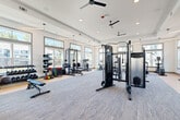 Located at 191 Commerce Dr in Villa Rica, Georgia, this fitness center offers a generously sized, single-floor workout facility designed to support a comprehensive range of exercise routines. The space features a clean, modern aesthetic with crisp white walls, a tray ceiling accented by recessed lighting and pendant fixtures, and multiple ceiling fans that promote air circulation throughout.

Large, multi-pane windows line the perimeter walls, flooding the interior with abundant natural light and providing views of the surrounding community. The flooring transitions between durable hardwood-style surfaces in the free weights area and plush, textured carpet in the central and cardio zones, creating distinct workout areas within the open layout.

The strength training section is well-appointed with a full rack of dumbbells, a barbell bench press setup, cable and pulley multi-station machines, and functional training accessories including medicine balls, kettlebells, and a plyo box. A dedicated stretching and mat area with interlocking foam tiles offers space for flexibility and cooldown work. The cardio zone features multiple treadmills, elliptical machines, stationary bikes, and a rowing machine, all positioned to take advantage of the natural light and outdoor views. Full-length mirrors enhance the sense of openness while providing visual feedback during workouts. The overall atmosphere is bright, airy, and purpose-built for fitness.