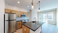 This contemporary single-floor residence showcases a thoughtfully designed open layout with light wood-tone flooring throughout, creating a warm and cohesive aesthetic. The kitchen is a standout feature, equipped with stainless steel appliances including a refrigerator, range, and over-the-range microwave, complemented by warm wood cabinetry, a classic subway tile backsplash, and deep dark granite countertops. A generously sized center island with a matching granite surface, an undermount sink, and a brushed gold faucet serves as the focal point, with stylish pendant lighting overhead. Sliding glass doors connect the living area to a private balcony, offering urban views and abundant natural light. The bedrooms are bright and comfortable, featuring light wood-tone flooring, large windows with city views, and ample closet space with sliding doors. The unit includes two bathrooms: one with a sleek walk-in shower finished in crisp white tile and a linear drain, and a second featuring a full soaking tub and shower combination with brushed nickel fixtures. Neutral wall tones and recessed lighting throughout contribute to a clean, modern ambiance well-suited for contemporary living.