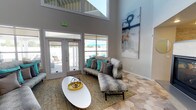 This single-floor community clubhouse is designed with a contemporary aesthetic and an open, airy atmosphere. The space is immediately distinguished by its soaring double-height ceilings, which amplify the sense of openness throughout. Large windows and glass-paneled doors flood the interior with abundant natural light, while offering direct views of and access to the outdoor pool and patio area beyond.

The main lounge area features a distinctive hexagonal-patterned tile floor that adds visual interest underfoot, complemented by plush sectional sofas and accent chairs in neutral tones accented with teal and charcoal throw pillows. A sleek, built-in two-sided fireplace serves as a striking architectural focal point, framed by a stone surround and a mounted flat-screen display above. Large-scale abstract artwork adorns the walls, enhancing the modern, curated feel of the space.

A built-in kitchenette with white cabinetry, mosaic tile backsplash, and stainless steel appliances provides functional amenity for residents. The entry area features a glass-walled room, ideal for private meetings or office use. Throughout, the design balances clean lines, warm textures, and vibrant accent colors to create a welcoming and versatile communal environment.