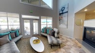 This single-floor community clubhouse is designed with a contemporary aesthetic and an open, airy atmosphere. The space is immediately distinguished by its soaring double-height ceilings, which amplify the sense of openness throughout. Large windows and glass-paneled doors flood the interior with abundant natural light, while offering direct views of and access to the outdoor pool and patio area beyond.

The main lounge area features a distinctive hexagonal-patterned tile floor that adds visual interest underfoot, complemented by plush sectional sofas and accent chairs in neutral tones accented with teal and charcoal throw pillows. A sleek, built-in two-sided fireplace serves as a striking architectural focal point, framed by a stone surround and a mounted flat-screen display above. Large-scale abstract artwork adorns the walls, enhancing the modern, curated feel of the space.

A built-in kitchenette with white cabinetry, mosaic tile backsplash, and stainless steel appliances provides functional amenity for residents. The entry area features a glass-walled room, ideal for private meetings or office use. Throughout, the design balances clean lines, warm textures, and vibrant accent colors to create a welcoming and versatile communal environment.
