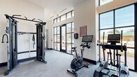This fitness center at 1407 Montgomery St in Savannah, GA, is a well-appointed, single-floor facility designed to support a wide variety of workout routines. The space is defined by its modern, open layout and an abundance of natural light, which streams in through large, black-framed floor-to-ceiling windows and clerestory windows above, offering street-level views of the surrounding Savannah neighborhood.

The facility is equipped with a thorough selection of cardio machines, including treadmills, an elliptical, and a stationary cycling bike—several of which feature integrated digital screens. A multi-functional cable machine serves as a centerpiece for strength training, while a dedicated free weights area includes a full rack of dumbbells, kettlebells, medicine balls, resistance bands, and a TRX suspension system. Additional strength machines and a weight bench further expand training options.

The interior features neutral white walls, dark baseboards, and textured gray carpet flooring that give the space a clean, contemporary aesthetic. A wall-mounted water fountain provides convenient hydration access. Fitness center rules are posted near the entrance, and ceiling fans help maintain airflow throughout. The overall ambiance is bright, functional, and well-organized, making it a practical and inviting space for daily exercise.