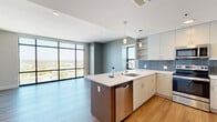 Located at 601 Bond Ave in Grand Rapids, MI, this modern two-bedroom, one-bathroom unit offers a thoughtfully designed open-concept layout with contemporary finishes throughout. The combined kitchen and living area serves as the heart of the home, featuring warm hardwood-style flooring that flows seamlessly across the entire unit. The kitchen is outfitted with sleek, light-toned cabinetry, a blue-gray tile backsplash, white quartz countertops, stainless steel appliances, and a central island with pendant lighting — ideal for both cooking and casual dining. Floor-to-ceiling windows in the living area flood the space with natural light while framing sweeping panoramic views of the Grand River, surrounding cityscape, and lush greenery beyond. The primary bedroom is a well-proportioned retreat with a large black-framed window offering elevated city and river views. The second bedroom features warm wood-tone flooring and clean neutral walls, providing a versatile space. The bathroom is finished with large-format tile surrounding the soaking tub and shower, a white vanity with quartz countertop, and chrome hardware. A dedicated closet completes the layout. The unit's modern, minimalist design aesthetic and elevated vantage point make it a standout residence in downtown Grand Rapids.