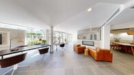 This single-floor space presents a cohesive modern aesthetic defined by clean lines, neutral tones, and thoughtfully curated furnishings throughout. The main lounge area is an open, light-filled space anchored by large floor-to-ceiling glass doors that invite abundant natural light and provide views of an outdoor area with lush greenery. Large-format gray tile flooring runs throughout, unifying the space visually. A seating arrangement of rich cognac leather chairs and sofas clusters around a sleek linear fireplace set into a travertine and mirrored tile surround, creating a refined focal point.

A dedicated work area features minimalist black desks and modern swivel chairs, positioned to take advantage of natural light from the expansive glazing. Adjacent to the main lounge, a dining area offers a round gray table surrounded by woven rattan chairs in warm amber tones, accented by a woven pendant light and abstract wall art. The double-sided fireplace shared between the lounge and dining areas adds a striking architectural detail.

The communal kitchen is well-appointed with light wood cabinetry, stone countertops, an integrated sink, and a patterned gray backsplash tile extending to open shelving. A media room rounds out the space, featuring a built-in flat-screen television, a sectional sofa, and curated artwork, offering an intimate retreat within this versatile, amenity-rich environment.