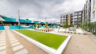 This thoughtfully designed outdoor amenity courtyard in Arvada, Colorado serves as a dynamic communal hub set against the backdrop of a modern multi-story residential building. The space is organized around several distinct activity and relaxation zones that flow seamlessly into one another. Synthetic turf bocce ball courts anchor the layout, providing a well-maintained recreational focal point framed by clean concrete borders. Adjacent to the courts, a covered pergola shelters an outdoor dining area furnished with bright teal chairs and tables, ideal for casual gatherings. A bold, large-scale mural in teal and white geometric patterns adorns the parking structure facade, adding a striking artistic element that energizes the space with color and visual interest. Thoughtfully curated xeriscape landscaping features native grasses, ornamental plantings, decomposed granite, and young deciduous trees that provide texture and greenery throughout. Concrete pathways connect each zone with smooth, accessible transitions. A fenced pet-friendly turf lawn offers a dedicated green area, while lounge seating positioned near the mural wall provides a relaxed social setting. The overall design balances functionality and aesthetics, creating a welcoming, community-oriented outdoor environment.
