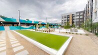 This thoughtfully designed outdoor amenity courtyard in Arvada, Colorado serves as a dynamic communal hub set against the backdrop of a modern multi-story residential building. The space is organized around several distinct activity and relaxation zones that flow seamlessly into one another. Synthetic turf bocce ball courts anchor the layout, providing a well-maintained recreational focal point framed by clean concrete borders. Adjacent to the courts, a covered pergola shelters an outdoor dining area furnished with bright teal chairs and tables, ideal for casual gatherings. A bold, large-scale mural in teal and white geometric patterns adorns the parking structure facade, adding a striking artistic element that energizes the space with color and visual interest. Thoughtfully curated xeriscape landscaping features native grasses, ornamental plantings, decomposed granite, and young deciduous trees that provide texture and greenery throughout. Concrete pathways connect each zone with smooth, accessible transitions. A fenced pet-friendly turf lawn offers a dedicated green area, while lounge seating positioned near the mural wall provides a relaxed social setting. The overall design balances functionality and aesthetics, creating a welcoming, community-oriented outdoor environment.