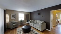 This single-floor space presents a thoughtfully designed living area that balances modern aesthetics with a comfortable, inviting atmosphere. The living room serves as the focal point of the space, anchored by a bold dark charcoal accent wall that creates a striking contrast against the surrounding warm taupe walls and bright white ceiling. Hardwood flooring runs throughout, adding warmth and a timeless quality to the room. Natural light filters in through a window, brightening the space and highlighting the clean lines of the interior. The room is furnished with a light gray sofa and a coordinating armchair, arranged around a round leather ottoman that doubles as a central coffee table. A patterned area rug in neutral tones grounds the seating arrangement and adds textural interest underfoot. Decorative touches include a framed wall mirror, wall art, and a small plant display on an ornamental side table, contributing to the overall polished and curated feel. A wall-mounted thermostat and visible HVAC vents indicate climate control throughout. The space reflects a cohesive, contemporary design sensibility with practical functionality and clean, understated elegance throughout its single level.