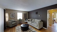 This single-floor space presents a thoughtfully designed living area that balances modern aesthetics with a comfortable, inviting atmosphere. The living room serves as the focal point of the space, anchored by a bold dark charcoal accent wall that creates a striking contrast against the surrounding warm taupe walls and bright white ceiling. Hardwood flooring runs throughout, adding warmth and a timeless quality to the room. Natural light filters in through a window, brightening the space and highlighting the clean lines of the interior. The room is furnished with a light gray sofa and a coordinating armchair, arranged around a round leather ottoman that doubles as a central coffee table. A patterned area rug in neutral tones grounds the seating arrangement and adds textural interest underfoot. Decorative touches include a framed wall mirror, wall art, and a small plant display on an ornamental side table, contributing to the overall polished and curated feel. A wall-mounted thermostat and visible HVAC vents indicate climate control throughout. The space reflects a cohesive, contemporary design sensibility with practical functionality and clean, understated elegance throughout its single level.