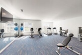 Located at 430 Mosby Blvd in Berryville, Virginia, this fitness center offers a well-equipped, single-floor workout facility designed to support a range of exercise routines. The space is laid out in a clean, open floor plan that maximizes usability and promotes easy movement between equipment stations.

The room features bright white walls and a smooth white ceiling with recessed and flush-mounted lighting, creating a well-lit environment throughout. Multi-colored, patterned carpet tiles in shades of blue, red, and gray cover the floor, adding a contemporary visual element while providing underfoot comfort during workouts.

A full-length wall mirror on one side of the room enhances the sense of space and allows users to monitor their form. Natural light filters in through several windows fitted with horizontal blinds, contributing to the open and airy atmosphere.

The fitness center is equipped with a diverse selection of cardio machines, including treadmills and stationary bikes — both upright and recumbent models. Strength training is well-represented with a cable/pulley multi-station machine, leg extension and curl machines, a weight bench, and dumbbell racks stocked with a full range of free weights. Exercise balls are neatly stored on a rack along the mirrored wall. Wall-mounted televisions provide entertainment, and an exit door offers direct access to the exterior of the building.