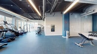 Located at 44 High St in Nashua, New Hampshire, this fitness center offers a generously sized, single-floor open layout designed to accommodate a full range of workout needs. The space features an industrial-inspired aesthetic, with exposed ductwork, steel piping, and bold linear LED lighting running along the dark ceiling, creating a modern and energetic atmosphere. Large windows along one wall flood the cardio zone with natural light and provide views of the surrounding Nashua streetscape, enhancing the workout experience.

The flooring throughout is a durable blue rubber material, well-suited for high-traffic fitness use. The cardio section is equipped with multiple treadmills, stationary bikes, and a stair climber. A dedicated strength training area features cable machines, a power rack, and a full dumbbell rack. A functional training zone includes adjustable benches, medicine balls, stability balls, resistance equipment, and wall-mounted mirrors to support form and technique. Ceiling fans contribute to ventilation and comfort throughout the space.

Storage cabinetry, a water station, and clearly marked emergency exits reflect thoughtful facility planning. Glass-panel partitions offer visual connectivity to adjacent areas. The overall design balances industrial character with a clean, practical layout suited for serious fitness use.