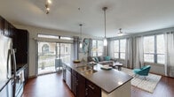 This single-story apartment in Spring, TX offers a thoughtfully designed open-concept layout that seamlessly connects the kitchen, dining, and living areas. The kitchen is a standout feature, equipped with dark espresso cabinetry, stainless steel appliances including a refrigerator, range, and microwave, granite-style countertops, and a large center island with a double sink — ideal for both cooking and casual dining. Pendant lighting and track lighting add functional elegance overhead. The living area is bright and inviting, with rich hardwood-style flooring, large windows on multiple walls that flood the space with natural light, and direct access to a private balcony with outdoor views. The bedroom offers plush carpeting, generous natural light through oversized windows, and a ceiling fan. The full bathroom features dark wood vanity cabinetry, a wide countertop with an undermount sink, a large mirror, and a tub/shower combination. A dedicated in-unit laundry area accommodates a full-sized washer and dryer with additional shelving for storage. The overall ambiance is modern and welcoming, with a cohesive design palette throughout.