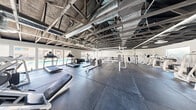 Located at 4650 W Oakey Blvd in Las Vegas, NV, this fitness center offers a generously sized, single-floor exercise room designed to accommodate a full range of workout activities. The space features a striking industrial aesthetic, defined by an exposed ceiling with visible steel trusses, wooden beams, and HVAC ductwork running throughout — lending the room a raw, warehouse-style character.

The flooring consists of large-format dark rubber tiles, providing a durable and impact-absorbing surface ideal for both heavy lifting and cardio training. The cardio zone is well-stocked with multiple treadmills and elliptical machines, positioned near floor-to-ceiling windows and glass doors that offer views of an outdoor pool area, allowing for natural light to fill the space.

The strength training area is equally comprehensive, featuring cable machines, multi-function cable crossover stations, a squat rack with barbell and weight plates, adjustable benches, and a full dumbbell rack with an extensive range of free weights. A dedicated selectorized machine section along one wall provides additional targeted muscle training options.

Horizontal windows near the roofline on the opposite wall bring in supplemental natural light, keeping the space bright and well-ventilated. The overall ambiance is functional yet modern, offering a complete fitness environment suited for all levels of training.