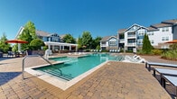 Located at 1004 Bailes Ridge Ave in Fort Mill, South Carolina, this two-story community property showcases a beautifully designed outdoor amenity space centered around a generously sized resort-style swimming pool. The pool features distinctive in-water sun shelves with modern loungers, allowing residents to relax while partially submerged, as well as traditional entry steps and handrails for easy access. Depth markers indicate both shallow and deeper sections, accommodating a range of swimmers.

Surrounding the pool is an expansive paver deck crafted from warm-toned stone, providing ample room for sunbathing and socializing. Rows of contemporary aqua-blue lounge chairs are neatly arranged along the pool's perimeter, offering abundant seating options. A white pergola structure anchors an outdoor dining and grilling area, complete with patio tables shaded by vibrant orange umbrellas, and a built-in grill set against a stone accent wall.

Mature trees and well-maintained landscaping with evergreen shrubs frame the space, providing natural privacy and shade. Classic black pole-mounted lanterns add a refined aesthetic detail throughout the deck area. The surrounding multi-story residential buildings, clad in blue and white craftsman-style siding with stone accents and private balconies, create an attractive and cohesive architectural backdrop. The overall ambiance is bright, open, and inviting under the clear Carolina sky.