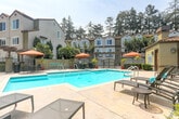 Located at 470 Saratoga Ave in San Jose, California, this outdoor amenity space showcases a striking Spanish Colonial-inspired design complemented by a generous, sparkling swimming pool as its central feature. The pool area is surrounded by a spacious travertine-style sundeck, thoughtfully furnished with cushioned lounge chairs and side tables, providing ample room for relaxation under the California sun. A separate spa is positioned adjacent to the main pool, offering a dedicated space for unwinding. Vibrant orange market umbrellas and potted plants add pops of color throughout the patio, enhancing the resort-like atmosphere. A well-appointed clubhouse building anchors one side of the space, featuring arched windows, clay tile roofing, and warm stucco exteriors consistent with the community's Mediterranean aesthetic. Tall, mature trees frame the perimeter, providing natural shade and a sense of privacy. Palm trees and lush tropical plantings reinforce the warm, Southern California-inspired ambiance. The entire area is enclosed by decorative fencing with gated access, ensuring a secure and intimate setting. The cohesive architectural style, abundant greenery, and carefully curated outdoor furnishings combine to create an inviting, community-focused retreat well-suited for leisurely gatherings and outdoor enjoyment.