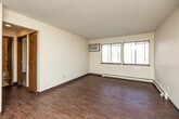 Located at 6316 Urbandale Ave in Des Moines, Iowa, this two-bedroom, one-bathroom unit offers a practical and functional single-floor layout. The living room is the heart of the space, featuring durable hardwood-style flooring, a textured popcorn ceiling, and a wall-mounted air conditioning unit. Large windows allow ample natural light to fill the room while offering views of the surrounding wooded scenery. Baseboard heating runs along the perimeter, providing warmth throughout the colder months. The kitchen, visible from the living area, includes warm wood-toned cabinetry and black appliances, offering a compact yet efficient workspace. Both bedrooms are carpeted for comfort and include windows with the same pleasant outdoor views. The primary bedroom provides a comfortable footprint with good natural light. The second bedroom, also carpeted, connects easily to the hallway for convenient access. The bathroom features a full tub-shower combination with classic beige tile surround and chrome fixtures, maintaining a clean and functional aesthetic. A connecting hallway with wood-toned doors ties all rooms together. This unit's efficient layout and consistent design details make it a straightforward, well-organized living space.