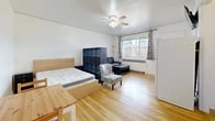Located at 1075 Myrtle St in Los Alamos, New Mexico, this single-floor studio unit offers a thoughtfully arranged open-plan layout that efficiently combines living, sleeping, and dining functions within a compact footprint. Warm-toned hardwood flooring runs throughout the main area, contributing to a bright and cohesive feel. A ceiling fan with integrated lighting is centrally positioned overhead, providing both airflow and illumination. Natural light enters through a large front-facing window dressed with patterned curtains, which also offers views of the surrounding neighborhood. A baseboard radiator beneath the window provides heating. The open living area accommodates a sleeping zone on one side, with the kitchen and dining area on the other, delineated by a freestanding shelving unit. The kitchen is equipped with a full-size refrigerator, a gas range, and warm wood cabinetry set against tile walls. A wall-mounted television is positioned for visibility across the main space. The bathroom features a standalone shower stall, a toilet, a vanity with sink, and tiled flooring. A closet with a coat hook panel near the front door provides additional storage. The unit's straightforward, functional design makes it a practical and self-contained living space.