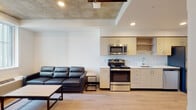 This single-floor unit showcases a contemporary design with a cohesive blend of industrial and modern elements. The open-plan living and kitchen area is immediately striking, featuring an exposed concrete ceiling with recessed lighting and a ceiling fan, set above light wood-style flooring that runs throughout the space. The living area accommodates a sectional leather sofa and a coffee table, with a window offering views of the surrounding urban streetscape, allowing natural light to fill the room.

The kitchen is efficiently laid out along one wall, equipped with a full suite of stainless steel appliances including a range, built-in microwave, dishwasher, and refrigerator. Light wood cabinetry with clean-lined hardware lines the upper and lower sections, complemented by white countertops and a single-basin sink. Open shelving adds both functionality and visual openness to the kitchen.

The bedroom is a well-proportioned space featuring the same exposed concrete ceiling, a ceiling fan with overhead lighting, and a window that provides natural light and exterior views. Built-in storage drawers beneath the bed frame maximize the room's utility. A dedicated workspace with a desk and chair completes the bedroom.

The bathroom is cleanly finished with a wide vanity topped in a stone-look surface, a large mirror, light wood cabinetry, and wood-tone flooring, maintaining the consistent modern aesthetic found throughout the unit.