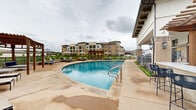 Located at 1400 N Main St in Keller, Texas, this resort-style outdoor pool area is a standout amenity within a well-appointed residential community. The centerpiece is a generously sized freeform swimming pool with clear, blue water and a smooth concrete pool deck that wraps around its curved perimeter, providing ample space for lounging and movement.

The pool deck is furnished with numerous cushioned chaise lounge chairs and patio seating, offering multiple relaxation zones. A rustic wooden pergola anchors one end of the pool area, providing a shaded retreat with additional seating beneath its open-beam structure. Umbrella-shaded tables are distributed throughout the deck, adding further comfort and sun protection.

Adjacent to the pool, a modern clubhouse building features brick and stucco exterior detailing, large glass windows and doors, and wall-mounted lantern lighting. Decorative hanging egg chairs flank the building's entrance, adding a stylish, casual touch. Manicured landscaping with trimmed shrubs and greenery lines the perimeter, contributing to a polished and inviting atmosphere.

The surrounding area offers open views of the broader community, including multi-story residential buildings visible in the background. A secured fence encloses the pool area, ensuring privacy and safety. The overall design balances functionality with resort-inspired aesthetics, creating an attractive and well-equipped outdoor recreational space.