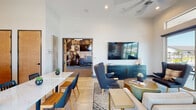 This single-story amenity space is thoughtfully designed to serve as a multifunctional community hub, seamlessly blending relaxation, fitness, and social dining into one cohesive environment. The open layout connects key areas fluidly, creating an inviting atmosphere throughout.

The lounge area anchors the space with soaring ceilings and generous natural light streaming through clerestory windows and large sliding glass doors. These doors open directly to an outdoor pool area, extending the living experience beyond the interior. The lounge is furnished with a plush neutral sofa, accent armchairs in deep blue tones, and a gold-accented coffee table ensemble, all arranged around a wall-mounted television atop a teal credenza. A ceiling fan overhead adds to the comfortable, casual ambiance.

Adjacent to the lounge, the kitchen is a standout feature, offering dark wood cabinetry, white quartz countertops, a subway tile backsplash, open wooden shelving, stainless steel appliances, and a large island with bar seating. Light tile flooring and high windows keep the space bright and airy.

The fitness center is equipped with treadmills, stationary bikes, and a wall-mounted television, with large windows providing ample natural light and outdoor views. Decorative acoustic panels and modern furnishings throughout contribute to the space's polished, contemporary aesthetic.