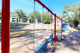 This outdoor recreational space offers a welcoming, open-air environment nestled within a residential community and surrounded by a rich canopy of mature trees. The grounds are characterized by a mix of gravel pathways and open grassy areas, creating a casual and accessible layout that invites exploration and leisure. Tall pines and deciduous trees provide natural shade throughout, casting dappled light across the terrain and contributing to a peaceful, park-like atmosphere.

A central feature of the space is a playground area equipped with a red swing set fitted with blue bucket-style seats, positioned on a ground cover of wood chips and packed earth that provides a safe surface for play. The playground is clearly visible and easily accessible from the surrounding open lawn, which gently slopes and offers ample room for outdoor activities.

Gravel paths wind through the property, bordered by landscape edging that helps define planting areas and pedestrian routes. In the background, multi-story residential buildings are visible through the tree line, situating this green space as a communal amenity within the broader neighborhood. A small structure or signage is also present near the tree line, suggesting organized use of the grounds. Overall, the space presents a functional and natural outdoor environment designed to serve the recreational needs of the surrounding community.