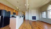 This one-of-a-kind loft conversion masterfully blends historic character with modern living. The open-concept living and kitchen area immediately impresses with soaring ceilings, original wide-plank hardwood floors, and a series of striking Gothic arched windows that flood the space with natural light and offer views of the surrounding cityscape. Exposed ductwork and structural elements add an industrial authenticity, while a built-in fireplace provides a refined focal point in the living area.

The kitchen is well-appointed with warm wood cabinetry, dark granite countertops, a tile backsplash, black appliances, and a central island with bar seating — ideal for both cooking and casual dining. Pendant lighting above the island adds an elegant finishing touch.

A long hallway connects the main living space to the private rooms, lined with multiple doors and flush-mounted lighting. One bedroom features charming beadboard wainscoting and a bright window, while another showcases an exposed brick accent wall with plush carpeting and an arched window — a nod to the building's storied past.

Two bathrooms serve the unit: one with dark slate tile flooring, a wood vanity, marble countertop, and a glass-enclosed shower; the other with light tile flooring, city-view windows, and a matching glass shower enclosure.