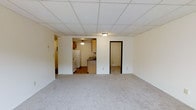 This single-floor space offers a functional and straightforward layout designed for practical everyday living. The main living area is finished with neutral-toned wall-to-wall carpeting, creating a comfortable and cohesive feel throughout. A drop tile ceiling with a central flush-mount light fixture provides even overhead illumination across the open space.

Visible from the living area, the kitchen features warm wooden cabinetry, a white refrigerator, a gas or electric range, and a sink set into a countertop with dark laminate detailing. The kitchen ceiling includes a pendant-style light fixture, adding a subtle distinction between the two areas. The kitchen flooring transitions to a harder surface, contrasting with the carpeted living space.

Two open doorways lead to additional areas of the unit. One doorway on the left appears to connect to a hallway, while the doorway on the right opens to a separate carpeted room, likely a bedroom. The overall color palette is neutral throughout, with white walls and ceilings contributing to a clean, modest aesthetic. The space presents a simple, utilitarian design suited for comfortable day-to-day use.