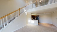This 1-bedroom, 1-bathroom residence with loft offers a well-organized two-story layout that balances open living with functional design. The main level centers around a spacious living area finished with plush neutral carpet and anchored by a striking staircase featuring a classic white spindle railing and warm wood handrail that draws the eye upward to the soaring double-height ceiling above. The open-concept kitchen is positioned adjacent to the living space and is outfitted with dark cabinetry, black appliances including a full-size refrigerator, gas range, and built-in microwave, along with a peninsula with a built-in sink that serves as a natural divide between cooking and living areas. Light tile flooring carries through the kitchen. The full bathroom on the main level features a tiled tub-shower combination, a dark wood vanity with a light countertop, a large mirror, and clean tile flooring. Upstairs, the loft level provides an open and versatile space with its own railing overlooking the living area below. The bedroom is a generously sized, carpet-floored room filled with natural light from a large window and is equipped with a ceiling fan. Additional windows throughout the residence bring in ample daylight, contributing to a bright, airy ambiance across both floors.