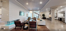 This well-appointed clubroom offers a sophisticated and inviting atmosphere designed for comfort and socializing. The space features an open-concept layout with distinct yet seamlessly connected lounge areas, each thoughtfully furnished to encourage both relaxed gatherings and casual conversation. A sleek wall-mounted flat-screen television is positioned above a fireplace framed by a textured white brick accent wall, adding warmth and visual interest to the main seating area. Rich, patterned area rugs anchor the furniture groupings, which include a plush white sofa accented with teal and gold throw pillows, deep red upholstered armchairs, and a dark wood table with coordinating seating. Overhead, a textured ceiling with recessed lighting and a central tray detail with ambient LED illumination adds architectural dimension to the room. A statement pendant light hangs above, complementing the layered lighting throughout. Large windows and glass doors at the far end draw in abundant natural light and provide views to an outdoor area, creating a bright and airy ambiance. Lush indoor plants enhance the space with a touch of greenery, while the overall design palette of neutral tones, warm woods, and bold accent colors conveys a polished yet approachable character.