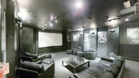 Located at 4130 Times Square Blvd in Dublin, Ohio, this single-floor private screening room delivers an immersive, cinema-quality experience within a thoughtfully designed entertainment space. The room is defined by its deep charcoal walls, dark ceiling, and recessed track lighting, creating a dramatic and intimate atmosphere reminiscent of a professional movie theater.

Dominating one wall is a large projection screen, positioned for optimal viewing from every seat in the room. Comfortable dark leather sofas and armchairs are arranged throughout the space, offering generous seating capacity for groups. A sleek glass-top coffee table anchors the central seating area, adding a polished, modern touch to the room's overall aesthetic.

Classic movie posters adorn the walls, reinforcing the cinematic theme and adding visual character to the dark-toned interior. The patterned carpet underfoot provides both comfort and acoustic benefits, contributing to the room's immersive sound environment. Subtle wall sconces complement the track lighting overhead, allowing for flexible ambiance control.

This space is purpose-built for entertainment, combining the comfort of a private lounge with the visual and atmospheric qualities of a dedicated screening room, making it an ideal venue for film screenings, presentations, or private events.