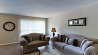 This single-floor space offers a warm and functional layout designed for everyday comfort. The living room serves as the heart of the space, featuring neutral wall tones and a textured ceiling that add subtle depth to the room. Large vertical-blind-covered windows allow generous natural light to filter in, creating a bright and airy atmosphere throughout the day. The room is furnished with two plush sofas accented with patterned throw pillows, arranged around a sleek glass-top coffee table, forming an inviting seating area ideal for relaxing or hosting guests. A classic table lamp provides warm ambient lighting, while a framed wall piece adds a decorative touch. The overall ambiance is calm and understated, with a cohesive neutral color palette of beige and grey tones that lend a clean, lived-in feel. The space strikes a balance between simplicity and comfort, making it a practical and welcoming environment.