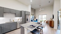 Located at 2911 E Sherman Dr in Denton, TX, this thoughtfully designed one-bedroom, one-bathroom residence offers a modern aesthetic with impressive architectural details throughout. The open-concept kitchen and living area is anchored by soaring ceilings that create an expansive, airy feel. The kitchen is outfitted with sleek gray shaker cabinetry, a white subway tile backsplash, granite countertops, stainless steel appliances, and a center island with bar seating — ideal for both cooking and casual dining. Light wood-style flooring runs throughout the main living area, complementing the clean, neutral palette. The living room is bathed in natural light from large sliding glass doors that open onto a private balcony overlooking lush greenery. The bedroom features plush carpet, tall ceilings, large windows with outdoor views, and access to two well-appointed closets with built-in shelving and hanging rods. The full bathroom includes a granite-topped vanity with gray cabinetry, a soaking tub with shower, and wood-style flooring. An in-unit laundry area adds everyday convenience. A smart home panel is also present, reflecting the unit's modern, tech-forward design throughout.