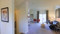 This two-story space showcases a thoughtfully designed interior with notable architectural character. The upper-level living area is a standout feature, boasting vaulted ceilings that lend an airy, open atmosphere to the room. Plush wall-to-wall carpeting runs throughout, adding warmth and comfort underfoot.

A sliding glass door floods the living area with abundant natural light while providing direct access to an outdoor area, framed by floor-to-ceiling patterned drapes that add a decorative touch. The living space is open and generously proportioned, accommodating a seating arrangement that includes modern swivel chairs in a bold orange tone, complemented by a low-profile coffee table.

A partial wall divides the space, creating a subtle sense of separation without compromising the open-plan feel. A slim shelving unit placed against this divider offers decorative display opportunities. Neutral white walls throughout keep the interior bright and versatile. Stairs connecting the two floors are visible, indicating a well-integrated multi-level layout. The overall ambiance is modern, light-filled, and welcoming.