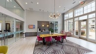 Located at 1200 Pickett Branch Rd in Cary, NC, this contemporary single-floor amenity space is thoughtfully designed to offer a wide range of communal features. The clubroom serves as the heart of the space, boasting soaring double-height ceilings, floor-to-ceiling windows that flood the area with natural light, and views of the surrounding landscape. A bold geometric-patterned area rug anchors a large wood dining table surrounded by vibrant magenta chairs, while a striking modern chandelier with globe bulbs adds an artistic focal point overhead. Glass-partitioned walls define a leasing or office area, maintaining visual openness throughout. A stylish kitchenette features gray subway tile backsplash, warm wood open shelving, quartz countertops, and stainless steel appliances. A separate lounge area with an energetic orange accent wall, plush seating, and a wall-mounted TV offers a comfortable retreat. The fitness center is expansive and well-appointed, featuring Life Fitness treadmills, Hammer Strength equipment, and colorful modular carpet tiles beneath vibrant teal accent walls. An adjacent spin and functional training studio provides space for cycling and group workouts, complete with exercise balls, resistance equipment, and mirrored walls. The overall design blends modern aesthetics with functional amenities.