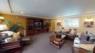 Located at 13011 Meridian E in Puyallup, Washington, this well-appointed lower-level space offers a generous, open layout designed for both relaxation and entertainment. The interior is finished with warm golden-tan accent walls complemented by crisp white trim and recessed lighting, creating a welcoming and polished atmosphere throughout.

The main living area serves as a dedicated media lounge, anchored by a large built-in dark wood entertainment center with glass-front cabinetry, housing a flat-screen television and ample storage. Plush wall-to-wall carpeting in a neutral tone runs throughout the space, adding comfort underfoot. Leather seating and rich wood furniture contribute to the room's warm, classic aesthetic.

A separate game area accommodates a full-size foosball table and connects seamlessly to the rest of the floor, making it ideal for entertaining. A staircase with a white-painted wood railing provides access between levels.

Multiple large windows allow natural light to filter in from multiple directions, keeping the space bright despite its lower-level position. Sconce lighting along the walls adds a layered, inviting glow. A reading nook corner with a leather armchair and ottoman sits beside a wide window bank, offering a quiet retreat within the broader entertainment space.