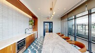 This stylish single-floor amenity space offers a thoughtfully designed layout spread across three distinct zones, blending bold interior design with seamless indoor-outdoor connectivity. The lounge area makes an immediate impression with deep teal-painted walls, a striking wood-slatted ceiling detail, and a vibrant mix of furnishings — including a mustard yellow sofa, a terracotta bench, and white lounge chairs — all anchored by a textured area rug. Built-in shelving with warm wood cabinetry and curated decor adds a refined, residential quality to the space.

The wet bar and kitchenette area is a standout feature, showcasing warm walnut cabinetry with brass hardware, dual sinks set into a veined white marble countertop, a subway-tiled backsplash with backlit floating shelves, and a stainless steel undercounter refrigerator. A geometric patterned teal floor tile flows throughout this zone, adding a playful yet sophisticated touch.

Adjacent to the kitchenette, a generous marble-topped island with leather barrel chairs serves as an ideal gathering or dining area, dramatically lit by a cascading brass pendant fixture. Floor-to-ceiling windows and sliding glass doors flood the space with natural light and open directly onto a furnished rooftop terrace, offering sweeping city skyline views.