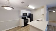 This single-floor residence offers a functional and well-organized layout with a clean, neutral aesthetic throughout. The open-concept kitchen and living area serves as the central hub of the space, featuring wood-look plank flooring that extends into the main living zone. The kitchen is equipped with white shaker-style cabinetry, white countertops, and a full suite of black appliances, including an electric range, over-the-range microwave, and refrigerator. A peninsula with an integrated sink and additional counter space connects the kitchen to the living area, providing both a practical workspace and a natural divider. The living room benefits from a ceiling fan with lighting and access to an outdoor area through sliding glass doors fitted with vertical blinds, allowing for natural light and ventilation.

The bedrooms are carpeted and similarly appointed, each featuring a ceiling fan with light fixture and a window with blinds that brings in natural light. The bathroom presents a streamlined design with a white vanity, white countertop, a large mirror, and wood-look flooring. A closet with louvered doors is also present in the unit. Throughout the space, a consistent color palette of soft gray and white tones contributes to a cohesive and understated ambiance.