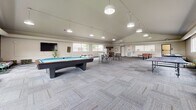 The community room at 1601 Belsly Blvd in Moorhead, Minnesota is a generously sized, single-floor gathering space designed to accommodate a variety of recreational and social activities. The room's most striking architectural feature is its vaulted ceiling, which rises prominently above the open floor plan and is lined with track-mounted pendant lights that provide even, welcoming illumination throughout. The flooring is a combination of neutral patterned carpet tiles and rich hardwood, delineating distinct activity zones within the space. Large windows along multiple walls flood the interior with natural light and offer views of the surrounding residential community. The room is equipped with multiple game tables, including a pool table, an air hockey table, and a ping pong table, making it well-suited for recreation. A variety of seating arrangements — from bar-height tables with stools to long dining-style tables with modern chairs and casual lounge seating — provide flexible options for gatherings of all sizes. A compact kitchenette with cabinetry and a sink supports event hosting needs. Navy blue accent walls add a bold design touch, complementing the otherwise neutral palette of white and gray throughout the space. A dedicated entry area leads directly into the main room, enhancing accessibility and flow.