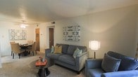 This single-floor residence offers a thoughtfully arranged living space designed for everyday comfort and functionality. The living room serves as the heart of the home, featuring plush carpeted flooring and a textured ceiling that adds subtle visual character. The space is furnished with a stylish gray sofa accented with decorative throw pillows in teal and gold tones, complemented by a sleek round side table and coordinating accent chair. A contemporary wall-mounted artwork adds a pop of color and personality to the neutral, light-colored walls. A floor lamp provides warm ambient lighting, enhancing the inviting atmosphere of the room. Visible from the living area, a dining space with seating is positioned to one side, creating a natural flow between entertaining and dining areas. Interior doors lead to additional rooms, suggesting a well-connected and practical floor plan. Overhead ventilation is integrated into the ceiling, ensuring climate comfort throughout. The overall ambiance is warm, approachable, and move-in ready, making this residence a comfortable and welcoming place to call home.